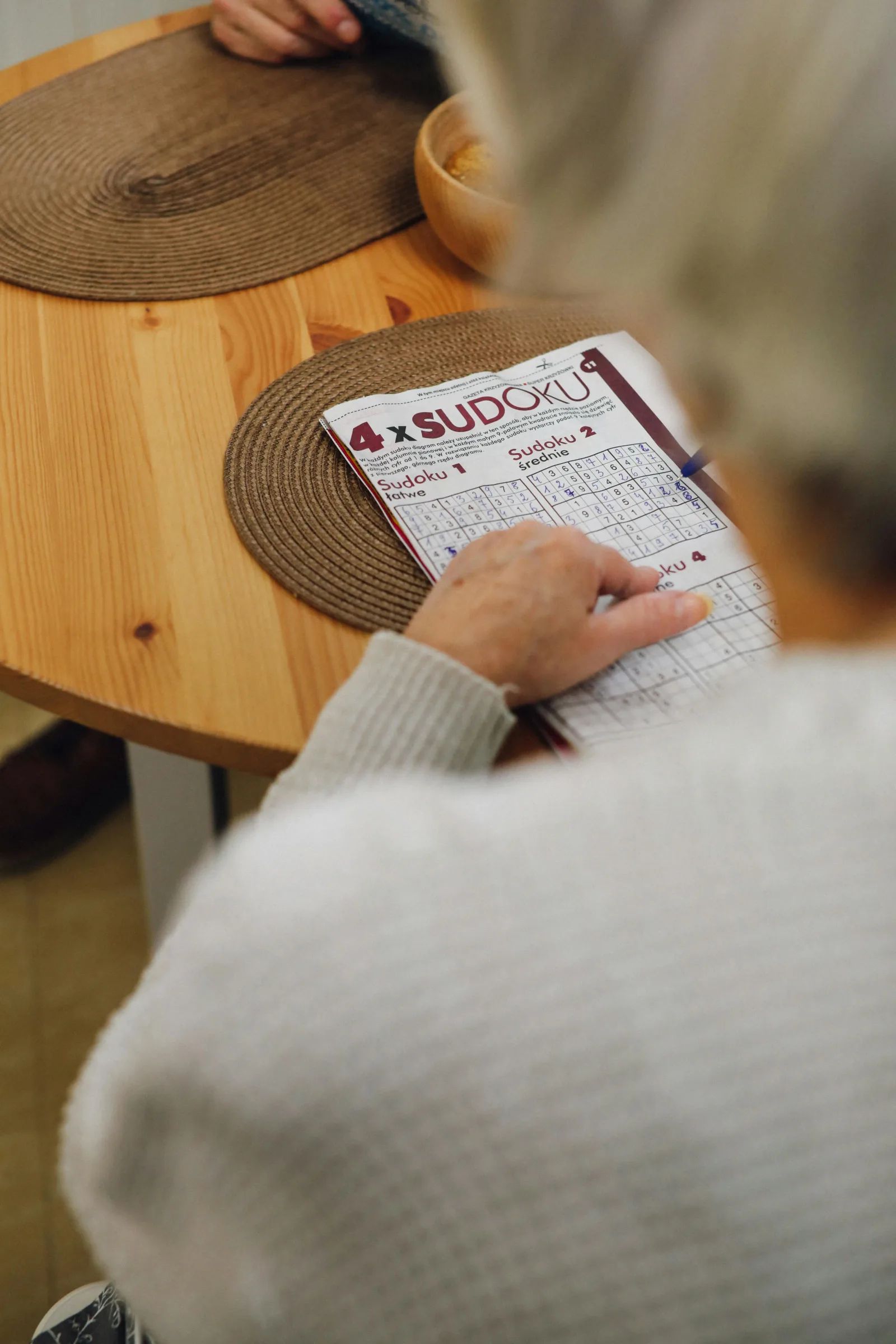 Vue par-dessus l’épaule d’une personne aux cheveux blancs qui remplit une grille de Sudoku dans un magazine posé sur une table en bois.