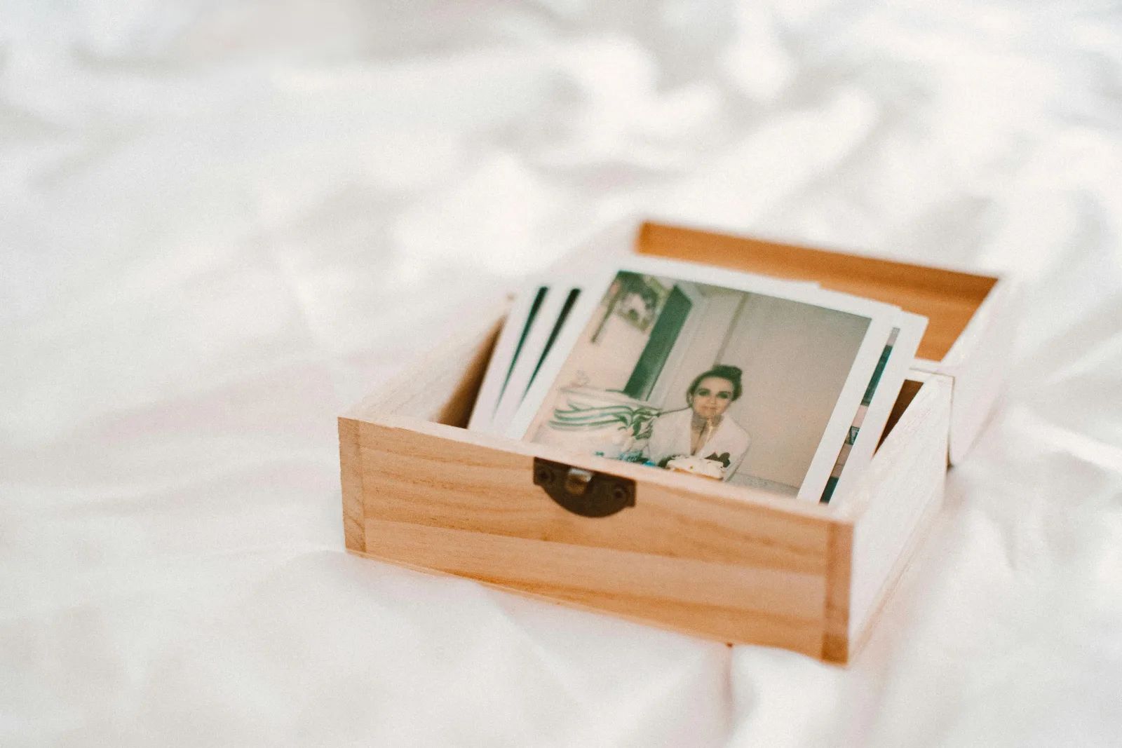 Une petite boîte en bois ouverte, posée sur un drap blanc, contient une pile de photos instantanées.