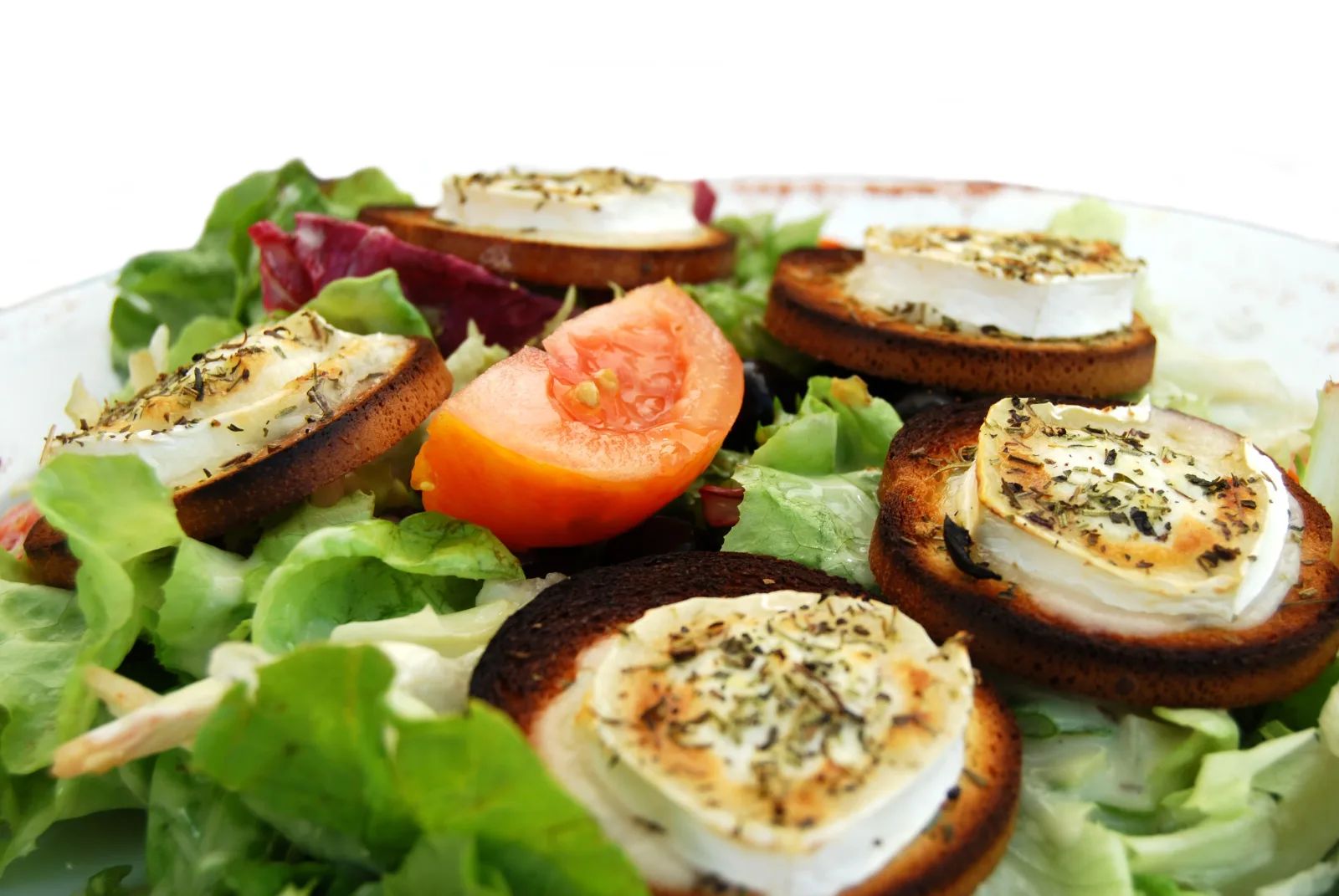 Une assiette de salade verte garnie de toasts de chèvre chaud aux herbes et d’un quartier de tomate.