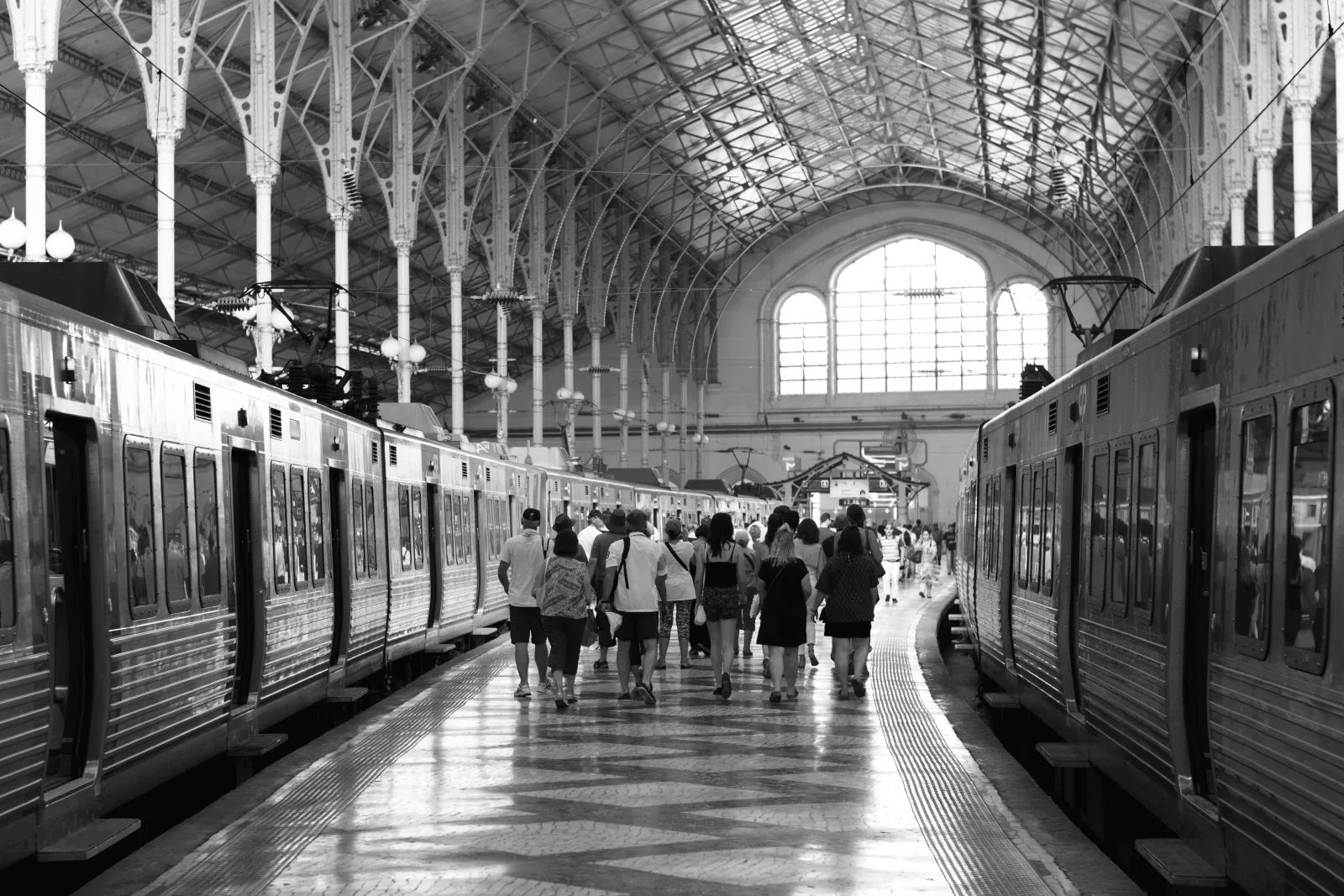 Photo en noir et blanc de voyageurs qui marchent sur le quai d’une gare, entre deux trains, sous une grande verrière.