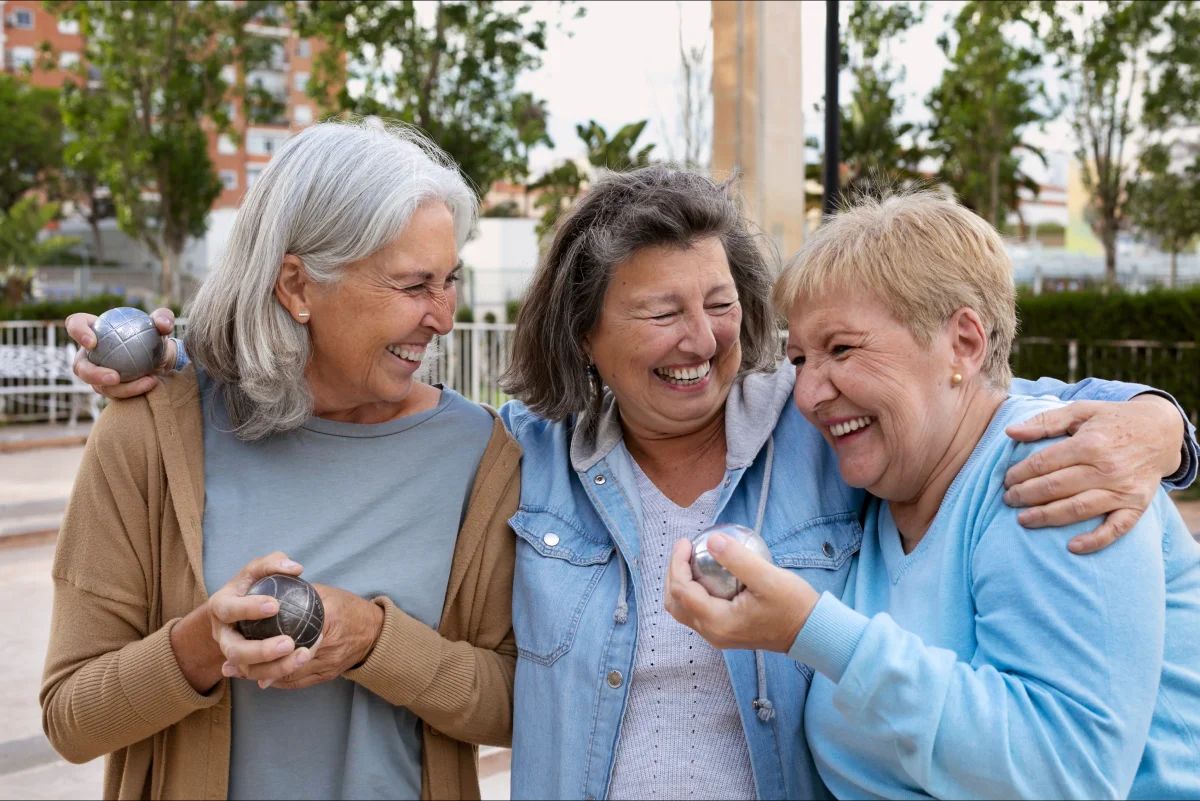 Trois femmes âgées rient en se tenant par les épaules, des boules de pétanque à la main.