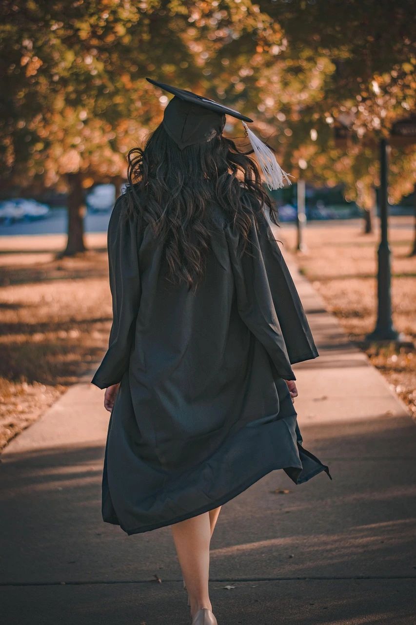 Une jeune diplômée, de dos, en toge et coiffe noires, s’éloigne sur une allée bordée d’arbres aux couleurs d’automne.
