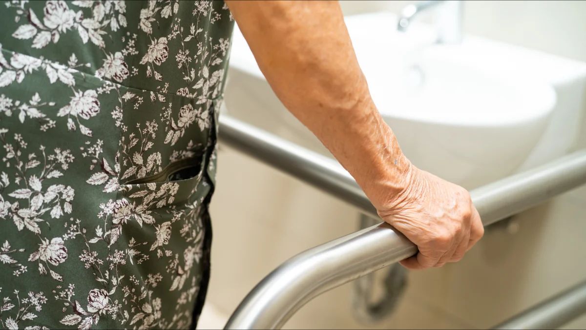 La main d’une personne âgée, vêtue d’une robe à fleurs, agrippe une barre d’appui en inox près d’un lavabo.