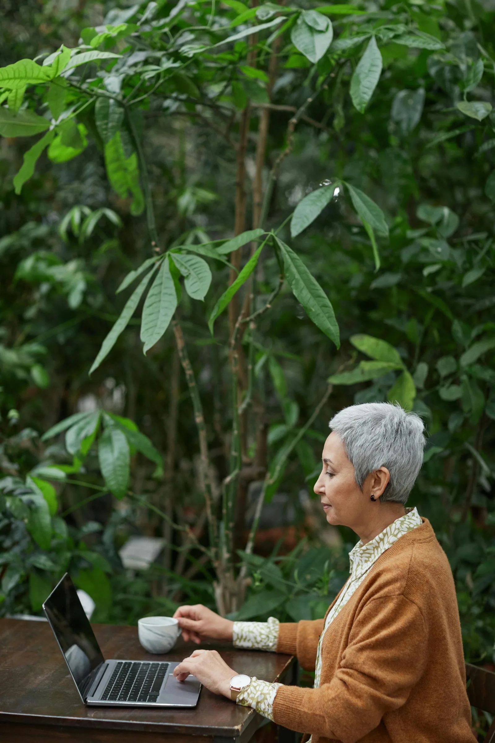 Une femme aux cheveux gris courts utilise un ordinateur portable, une tasse à la main, devant des plantes vertes.