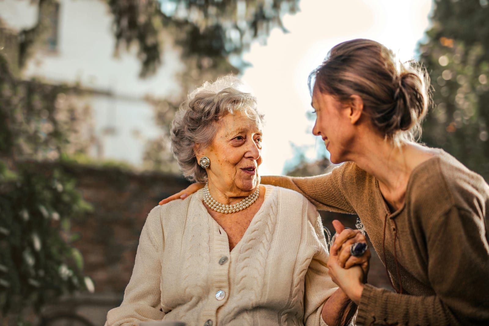 Une femme âgée en gilet clair et collier de perles échange un sourire avec une femme plus jeune qui lui tient la main, dans un jardin.