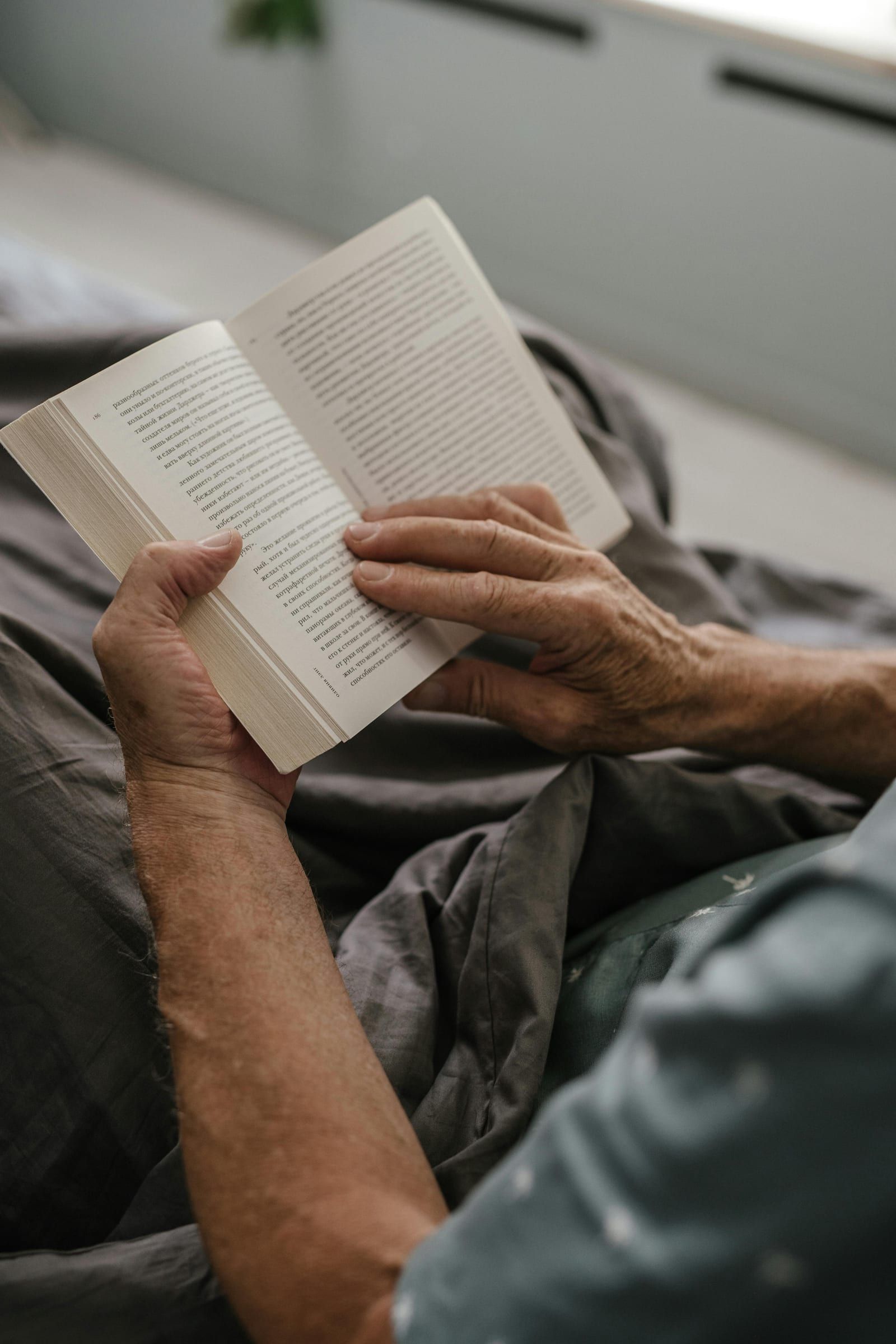 Les mains d’une personne âgée tiennent un livre ouvert, au creux d’une couverture grise.