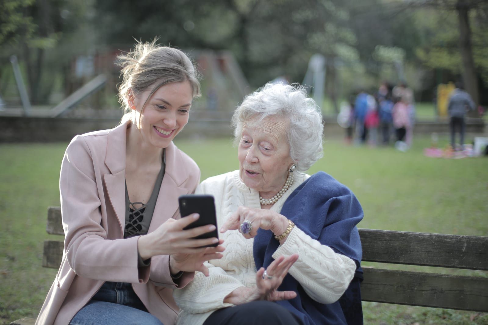 Une jeune femme montre l’écran de son téléphone à une dame âgée, assises côte à côte sur un banc dans un parc.