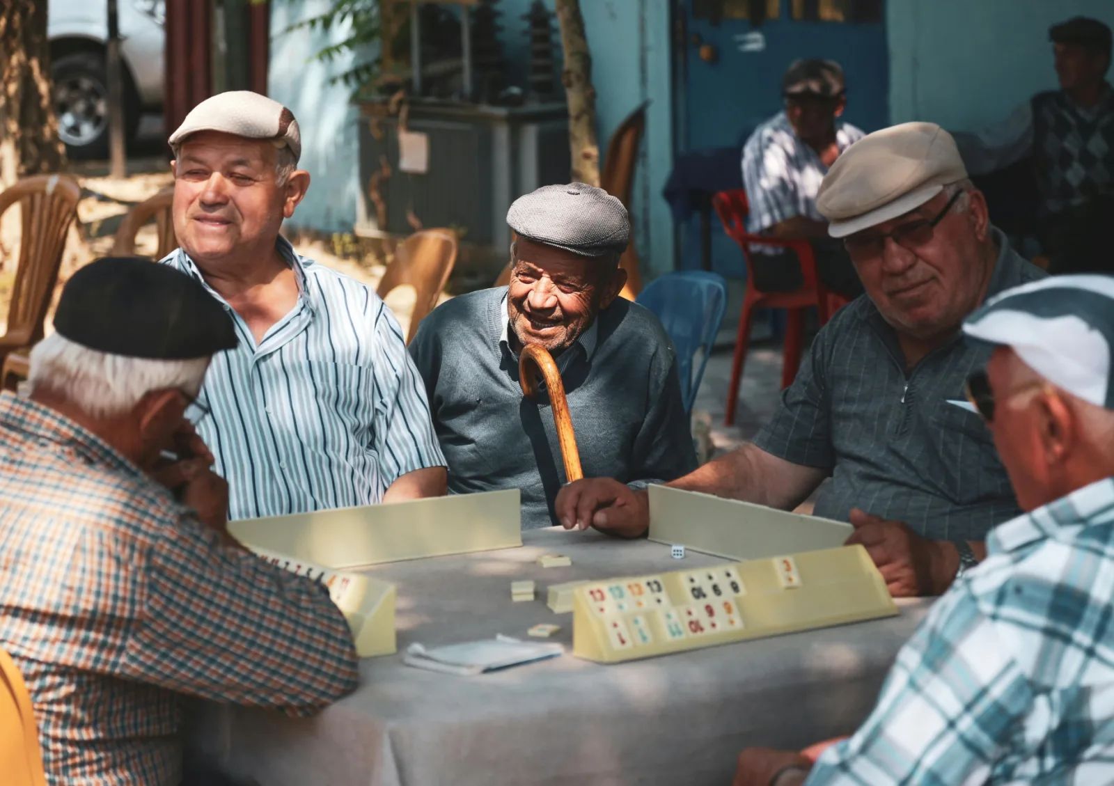 Des hommes âgés coiffés de casquettes jouent au rami autour d’une table, à l’ombre, et échangent des sourires.