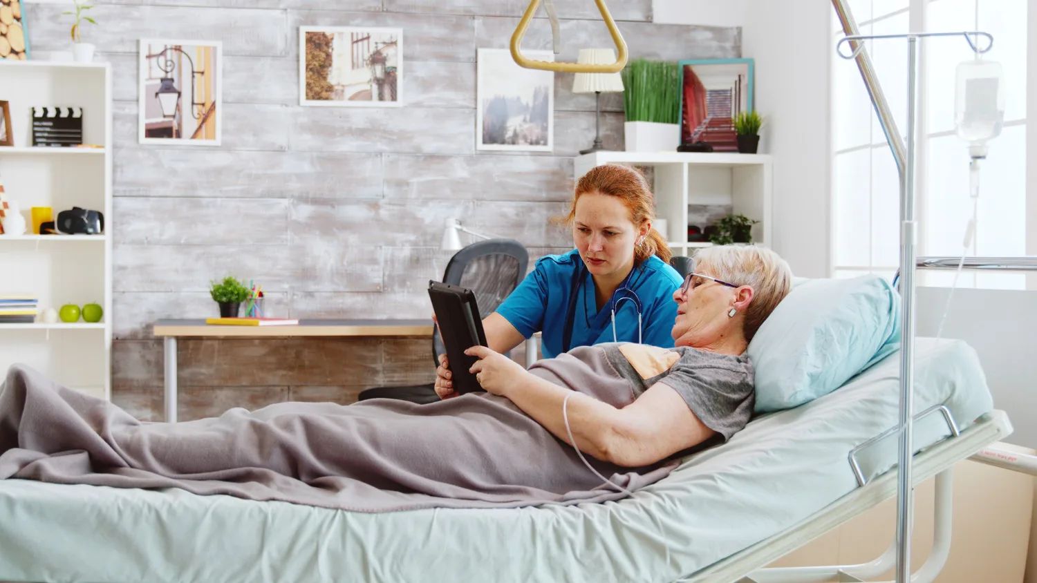 Une soignante en blouse bleue tient une tablette devant une femme âgée allongée dans un lit médicalisé, sous une potence.