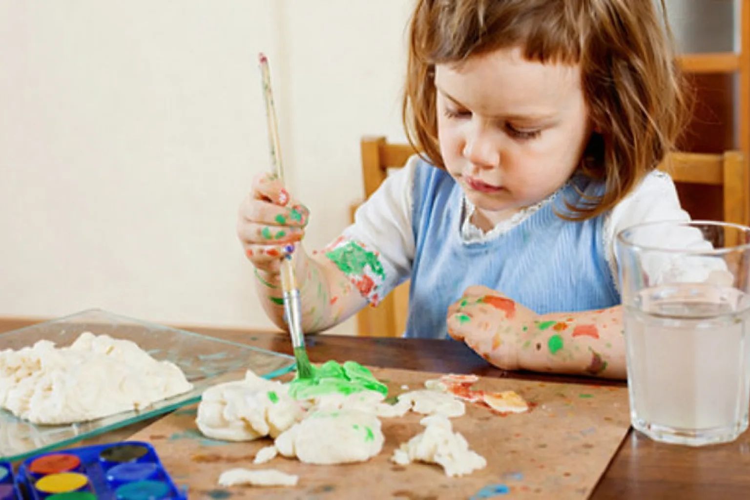 Une petite fille, les mains et les bras tachés de peinture, peint au pinceau des formes en pâte à sel sur une table.