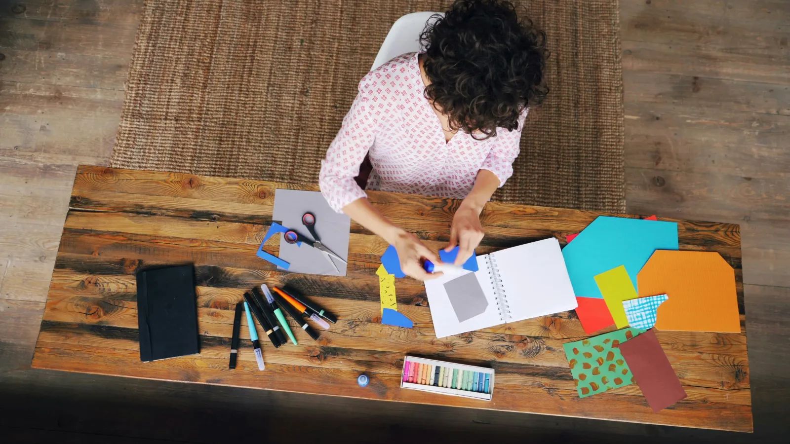 Vue de dessus : une femme colle des papiers de couleur dans un carnet, entourée de ciseaux, de feutres et de pastels.