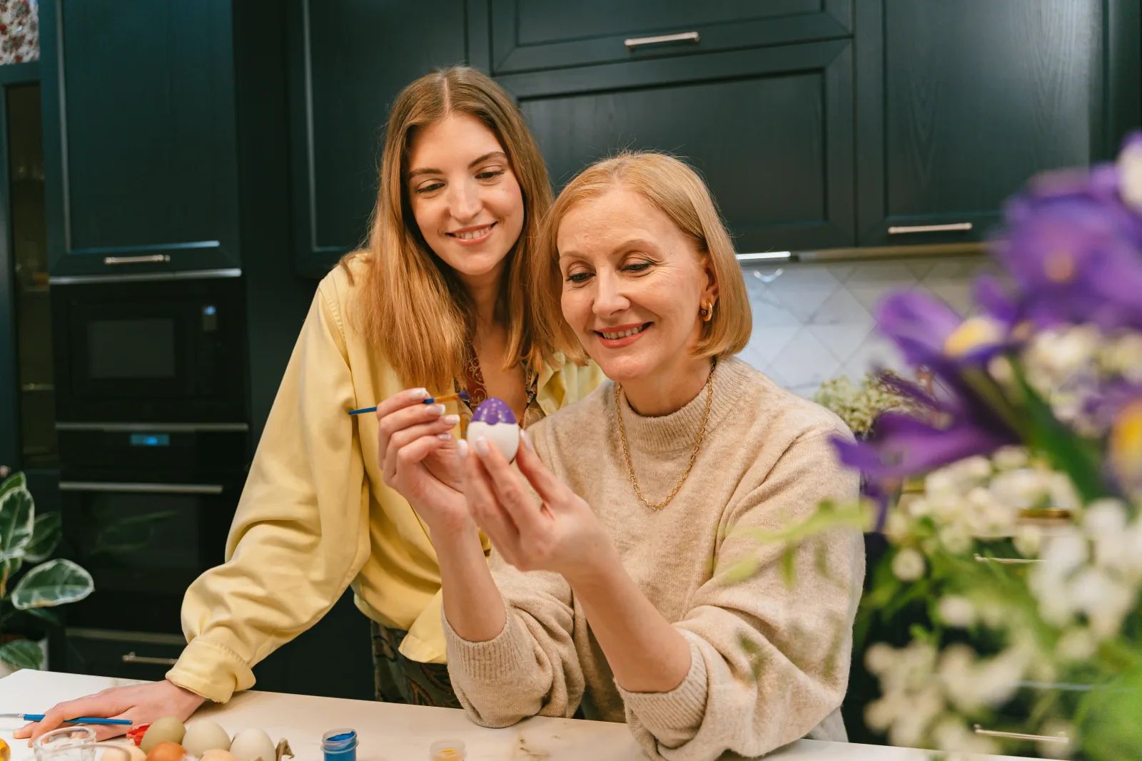 Deux femmes souriantes, l’une jeune, l’autre plus âgée, peignent un œuf de Pâques dans une cuisine, derrière un bouquet de fleurs.