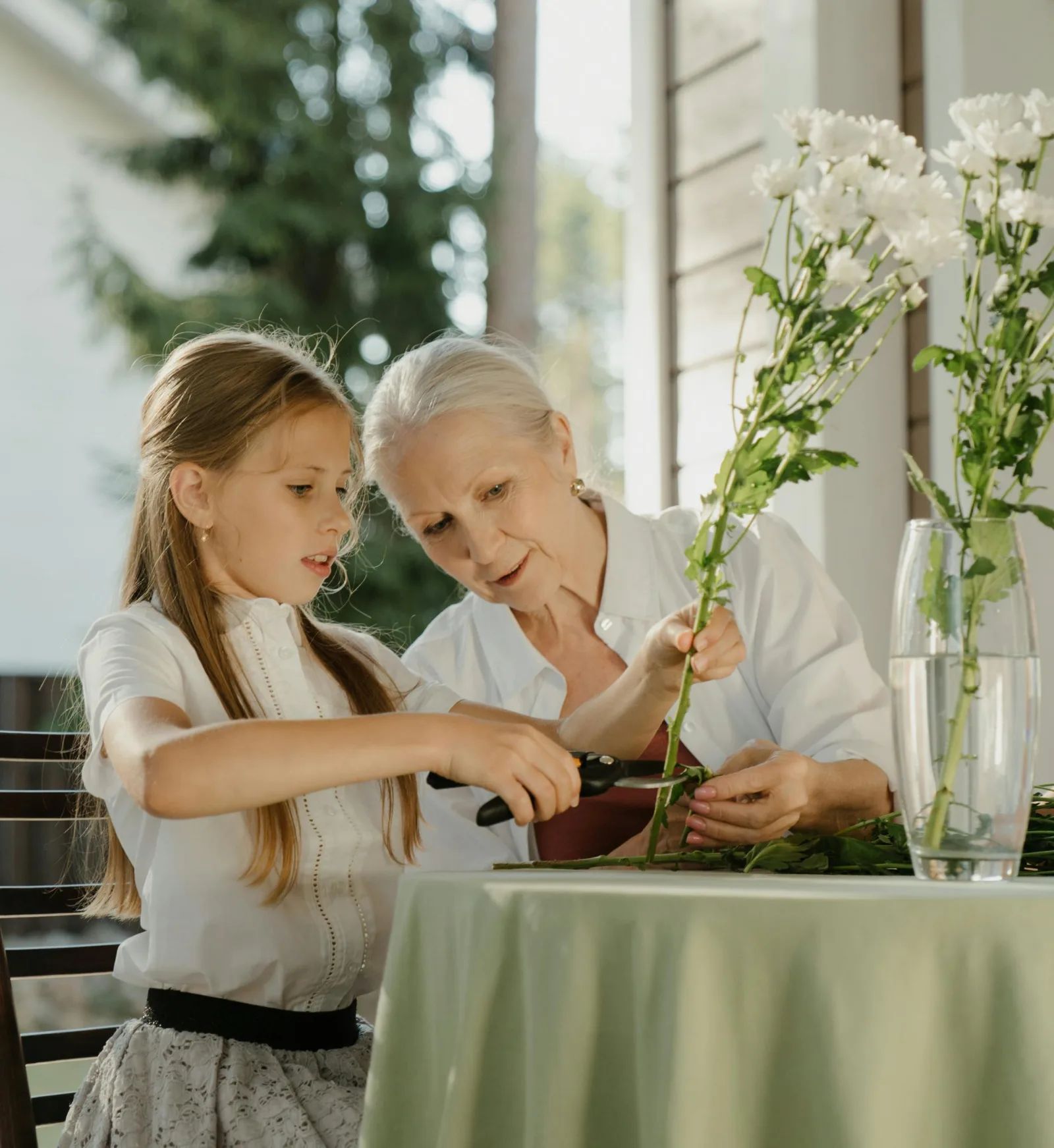 Une grand-mère et sa petite-fille, assises à une table, coupent la tige d’une fleur blanche pour composer un bouquet.