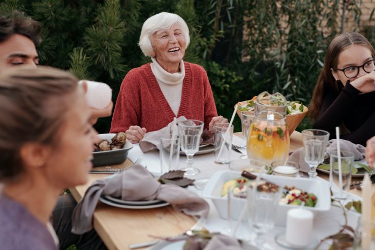 Une femme âgée en gilet rouge rit, entourée de convives, autour d’une table dressée dans un jardin.