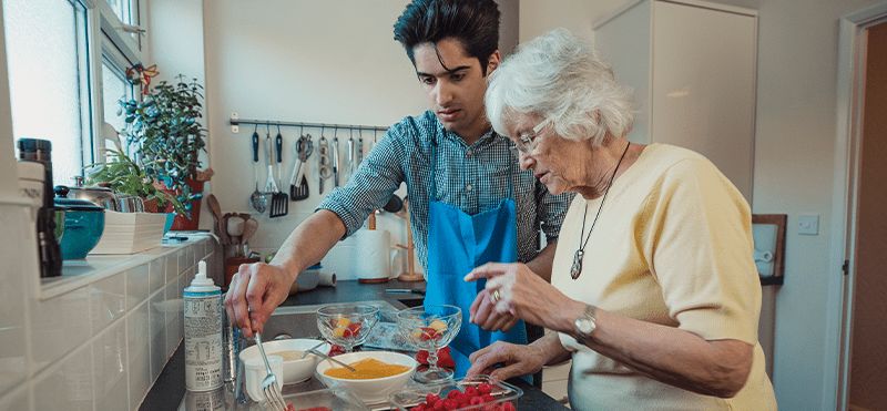 Une femme âgée et un jeune homme en tablier bleu préparent ensemble un dessert aux framboises dans une cuisine.